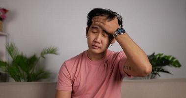 Portrait of Young Asian man getting cold and coughing while sitting on couch in living room at home. Unhappy Male coughs and covers his mouth using his hands. Common symptoms of Covid-19. video