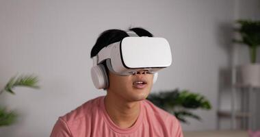 Portrait front view of a Young Asian man sitting on a sofa in the living room wearing a VR headset. Male having VR headset with many emotions, waving their hands in the air. Virtual reality technology video