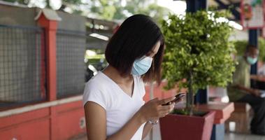 mulher asiática jovem viajante falando no celular e sentado no banco enquanto espera o trem na estação de trem. fêmea usando máscaras protetoras, durante a emergência covid-19. video