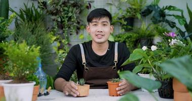 retrato de un joven jardinero asiático feliz que vende en línea en las redes sociales y mira la cámara en el jardín. hombre en videollamada con máscara facial. vegetación doméstica, venta en línea y concepto de hobby video