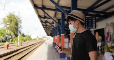 vista lateral do homem jovem viajante asiático andando e olhando um relógio na estação de trem. homem usando smartphone. macho usando máscaras protetoras, durante a emergência covid-19. conceito de transporte e viagens. video