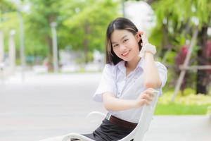 retrato de un estudiante tailandés adulto con uniforme de estudiante universitario. hermosa chica asiática sentada sonriendo felizmente en la universidad al aire libre con un fondo de árboles de jardín al aire libre. foto