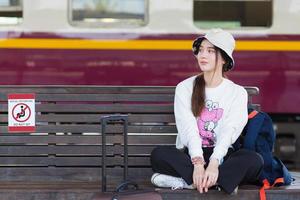 Asian woman in white long sleeve and hat sits on chair with her bag at train platform in social distancing as a new normal lifestyle. photo