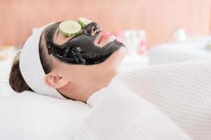 una hermosa mujer asiática usa barro de spa y pepino para el tratamiento facial foto