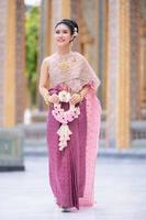 una hermosa y elegante mujer tailandesa vestida de tailandesa adornada con preciosos puestos de joyería sosteniendo una guirnalda de flores