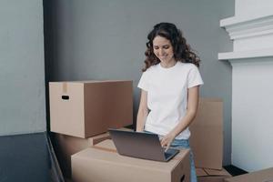 Happy european woman unloading cardboard boxes at new place and typing on laptop on the box.