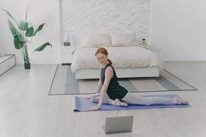 Red haired woman practicing yoga by video lessons in bedroom. Internet learning and home classes.