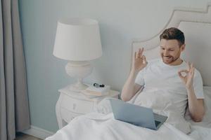 Happy positive young man sitting in cozy bed with laptop and enjoying video call photo
