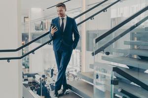 Stunning entrepreneur in formal stylish suit standing on staircase in office lobby with smartphone photo
