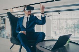 Well-groomed CEO man in formal suit wearing VR headset goggles, while sitting in office lobby