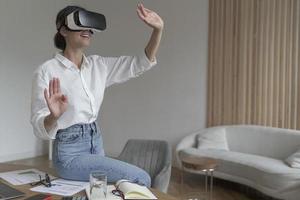 Joyful young business woman sitting on top of desk at home wearing VR glasses