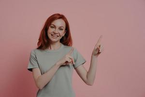 Satisfied pleased young redhead woman promoting product, pointing with index fingers at copy space