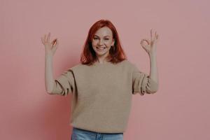 Half length shot of pretty ginger woman showing OK sign with both hands, isolated on pink photo