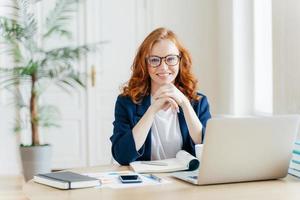 Working concept. Intelligent manager with satisfied expression, writes in notepad, works on modern laptop computer, uses organizer app, sits against office interior. Experienced entrepreneur photo