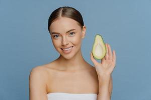 Natural skin care concept. Beautiful brunette young woman holds half of avocado enjoys organic cosmetics enjoys essential oils for beauty care poses wrapped in bath towel against blue background photo