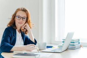 toma horizontal de la exitosa y confiada propietaria de una empresa pelirroja sentada en el gabinete, trabaja en una computadora portátil, escribe tesis principales en un cuaderno, tiene un descanso para tomar café, posa en una espaciosa sala de oficina foto