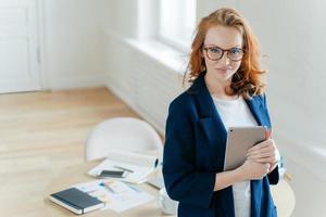 Shot of beautiful red haired young woman checks notification on touch pad, happy to recieve income message, looks confidently at camera, workplace with paper documents and notepad in background. photo