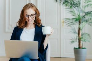 Satisfied European female with ginger hair, types information on laptop computer, has glad facial expression, drinks coffee, wears spectacles, develops new project, poses in cosy room alone. photo