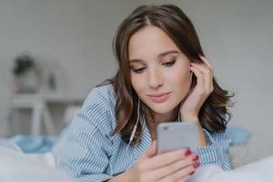 Shot of pretty brunette student learns language via mobile app, listens audio with earphones, has free time, dressed in casual outfit, poses on comfortable white bed, has thoughtful look aside photo