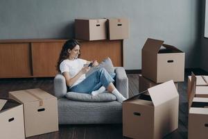 mujer en sillón hace clic en el teléfono entre cajas. chica europea descansando en el apartamento después de la reubicación. foto