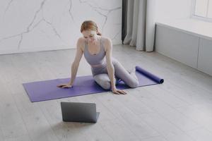 Young happy white woman practicing yoga watching video on laptop. Concept of e-learning. photo