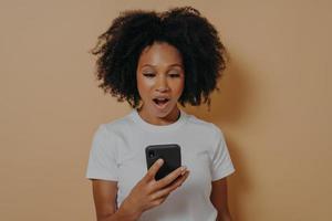 Surprised african female looking at smartphone with shocked face expression on beige background