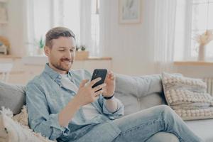 Happy smiling bearded man using smartphone device while relaxing on sofa photo
