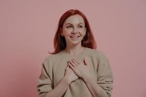 Young happy red-haired woman putting hands on chest while standing isolated on pink background