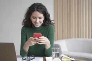 Young italian freelancer lady with dark wavy hair holding cell phone and looking at screen with smile