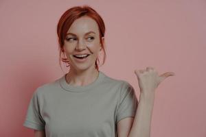 Amazed excited ginger female shop dressed in casual t-shirt pointing indicating with thumb photo