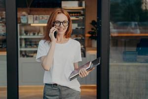 Smiling woman with laptop and notebook in hand enjoying talk on cellphone near coffee shop photo