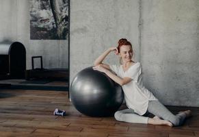 Cute sportive red haired female sitting on wooden floor with big silver fitball