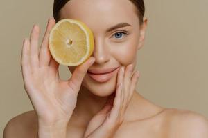 Cropped shot of tender good looking woman holds slice of fresh lemon over eye recommends organic cosmetics undergoes beauty procedures stands bare shouldered indoor. Natural treatment concept photo