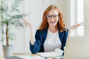 una foto de una mujer independiente positiva de cabello rojo trabaja de forma remota con documentos en papel, extiende las manos para mostrar su buen resultado, se sienta en el escritorio con un bloc de notas y una computadora portátil, prepara el trabajo del curso