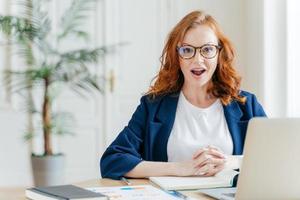 Surprised ginger female employee has job, develops new business srategy, poses in front of opened laptop computer, poses in workspace against office interior. Woman software developer at work. photo