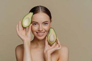 Facial treatment with natural vitamins. Pleased brunette woman smiles gently holds halves of avocado looks directly at camera stands topless indoor has healthy skin isolated over beige wall. photo