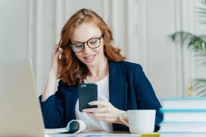 Photo of female freelancer uses smart phone for online communication, has red curly hair, wears optical glasses and formal costume, has coffee break after working on laptop computer, uses free wifi