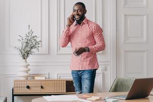 apuesto joven hombre de negocios africano exitoso en ropa informal disfrutando de una llamada telefónica, usando un teléfono inteligente foto