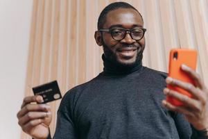 Cheerful African american businessman holding credit card and smartphone for shopping in internet photo