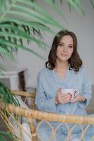 Adorable young female has charming look, dressed in striped domestic costume, drinks hot beverage, sits in wicker chair, enjoys rest and day off. People, lifestyle and recreation time concept photo