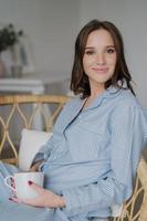 Satisfied beautiful woman with pleasant appearance, wears striped nightwear, drinks aromatic coffee, poses indoor. Relaxed housewife has drink at morning after awakening. People and rest concept photo