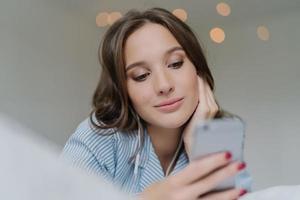 Shot of pretty brunette student learns language via mobile app, listens audio with earphones, has free time, dressed in casual outfit, poses on comfortable white bed, has thoughtful look aside photo