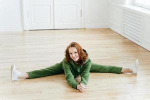 Horizontal shot of positive redhaired woman does pilates exercises, drssed in green tracksuit, has nice flexibility, practices yoga, looks at camera. People, active lifestyle and gymnastics concept photo