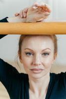 Headshot of talented skilled female ballet dancer poses near barre, has blue eyes, healthy skin, appealing appearance, practices dance in dancing hall. Red haired choreography instructor indoor photo