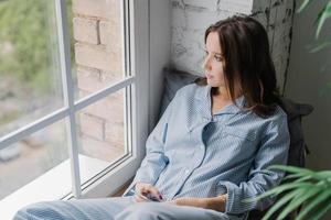 Horizontal shot of thoughtful young woman sits on windowsill, dressed in casual nightwear, uses smart phone and earphones, listens calm music while looks thoughtfully in window, enjoys home time photo