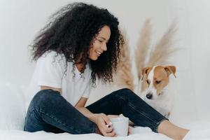 Photo of lovely curly haired female dog owner looks with smile at animal, holds cup of tea, sits on bed in white spacious bedroom, expresses love to animal. People, pets and friendship concept
