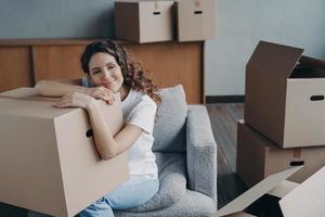 Hispanic girl relocates to new home. Lady is sitting in armchair among boxes and dreaming. photo