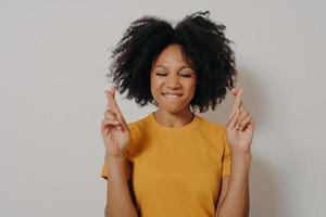Portrait of pretty cheerful african girl standing in studio and holding fingers crossed for luck photo