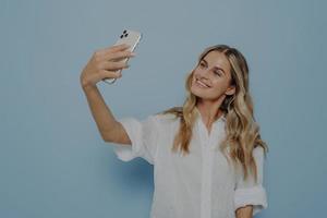 Beautiful young female with long blonde dyed hair holding mobile phone and posing for selfie photo