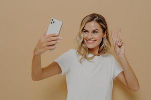 Cool joyful woman with blonde hair with smartphone in hand making selfie in studio against nude background